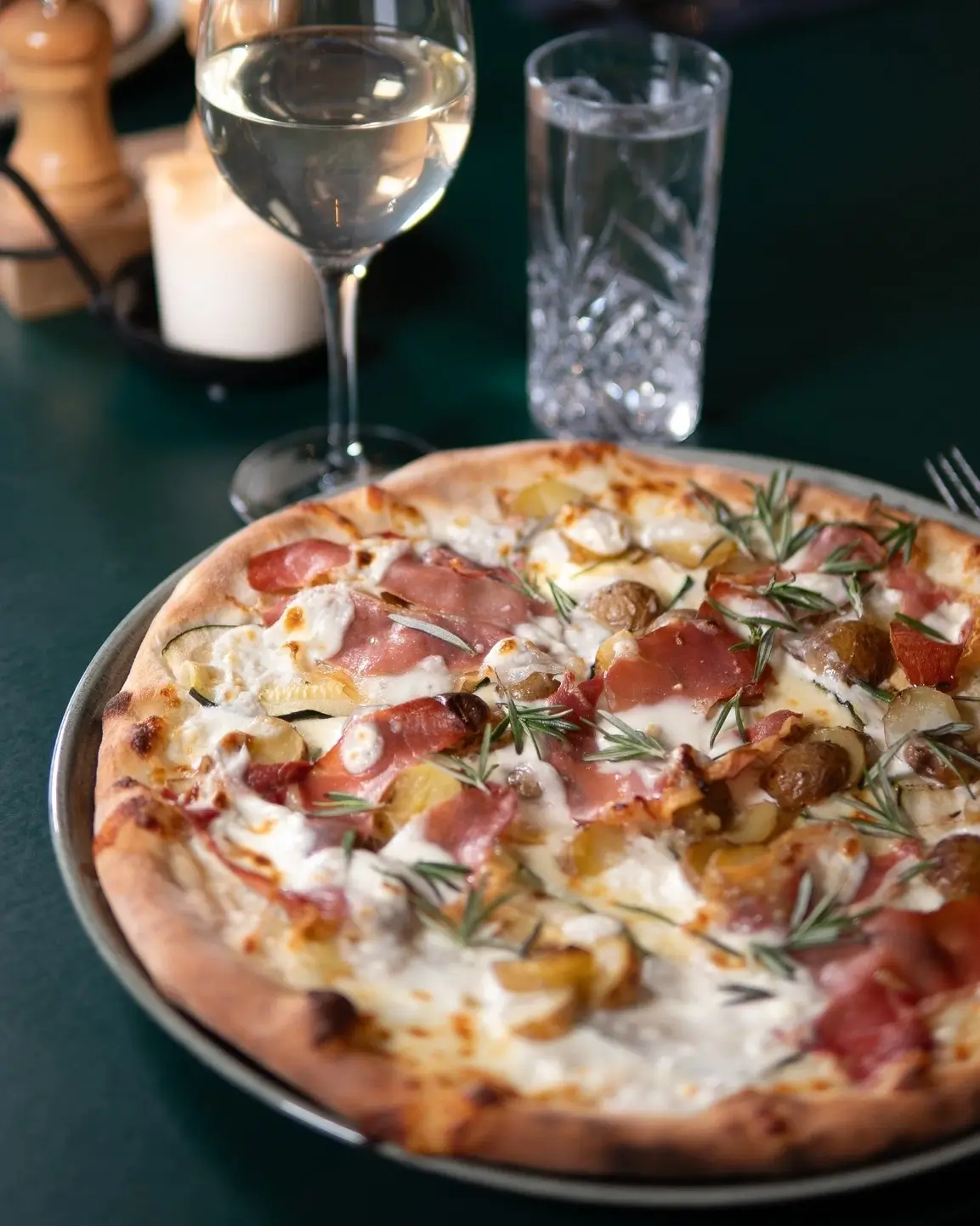 Pizza toppet med bacon, skiver af kartoffel, frisk rosmarin og smeltet ost, serveret med hvidvin og stearinlys.
